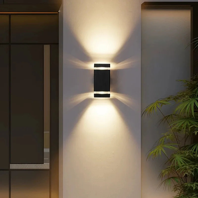 Modern wall-mounted outdoor light fixture on a building exterior with plants in the background.