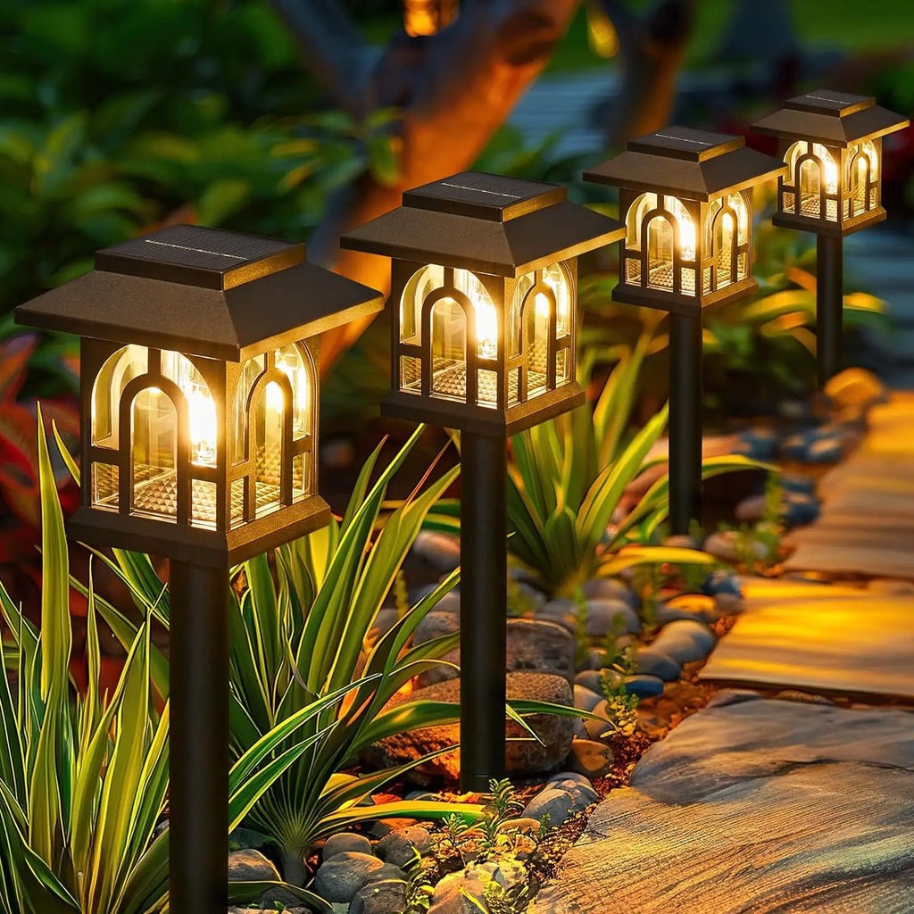 Set of four solar garden lights on a pathway surrounded by plants and rocks.