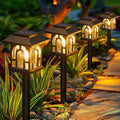 Set of four solar garden lights on a pathway surrounded by plants and rocks.